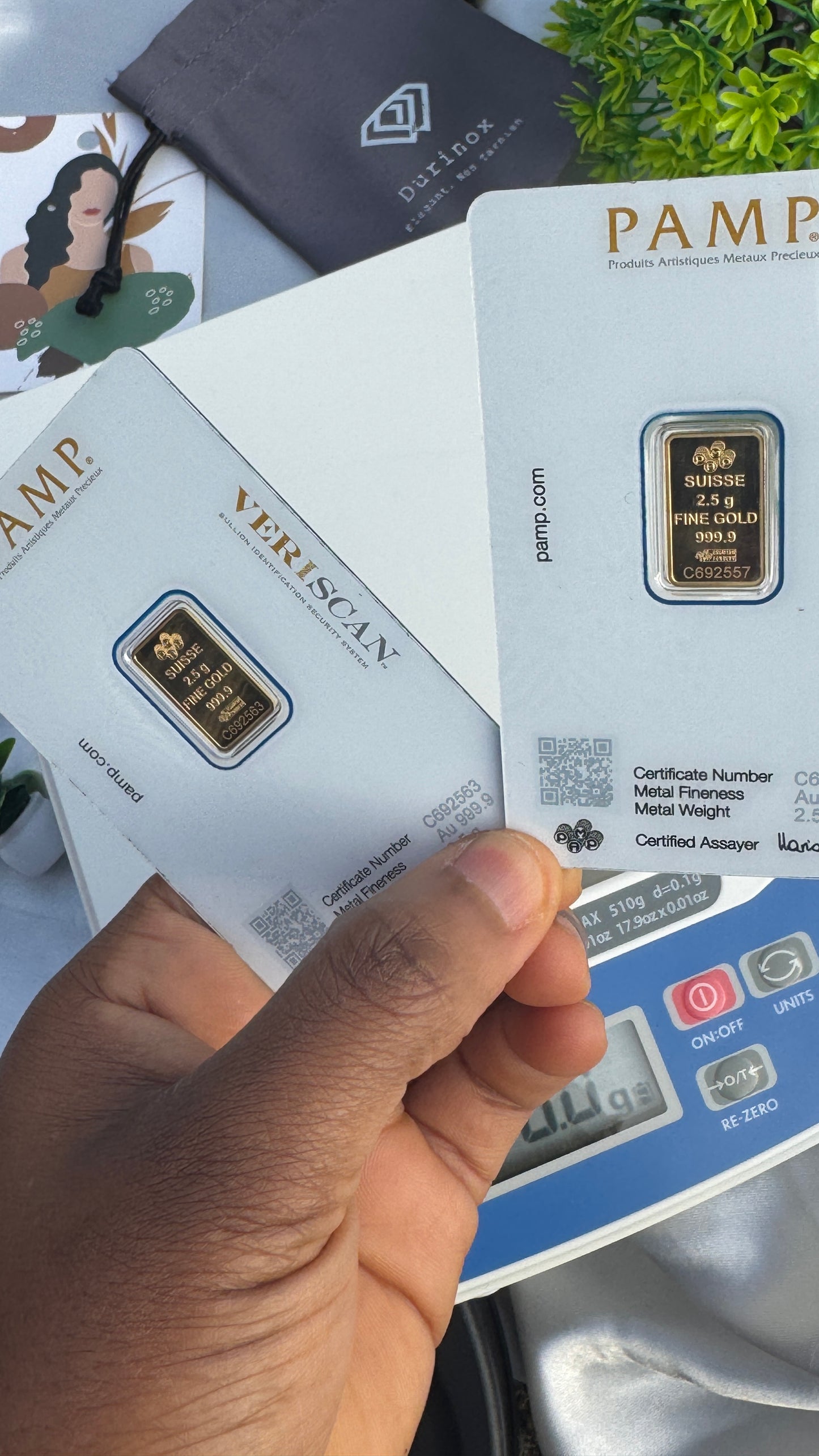 Hand holding a PAMP gold bar with packaging and a digital scale in the background