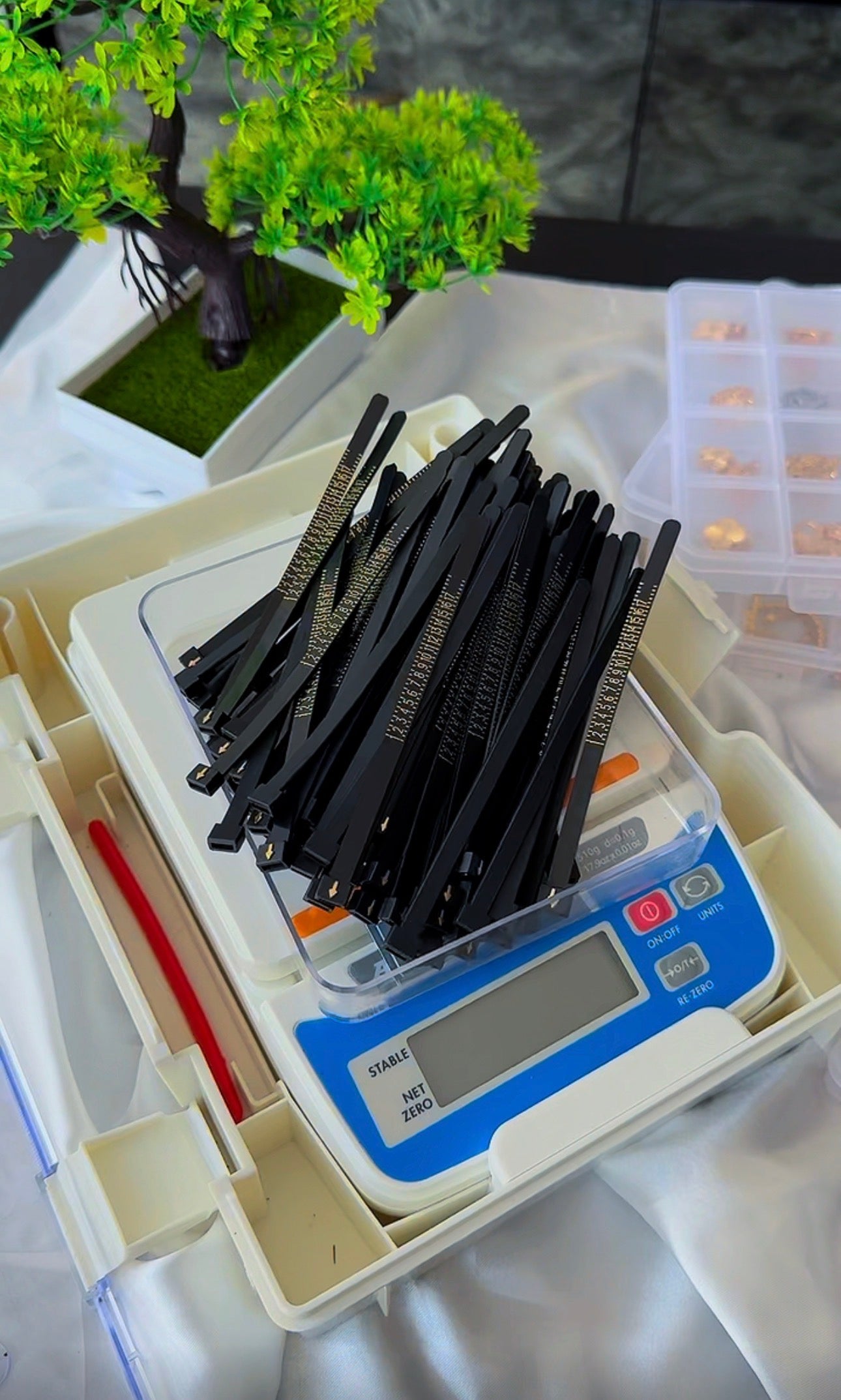 A tray of black hair extension strips placed on a digital scale, with a small potted plant and a plastic organizer visible in the background.