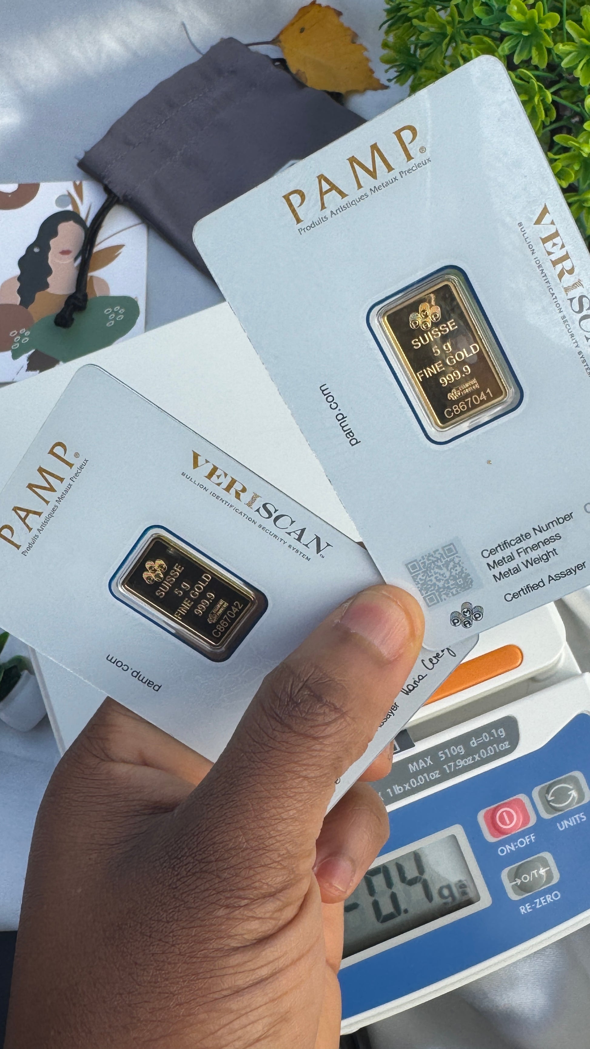 Hand holding a gold bar with 'PAMP' branding against a background of similar bars and a digital scale.