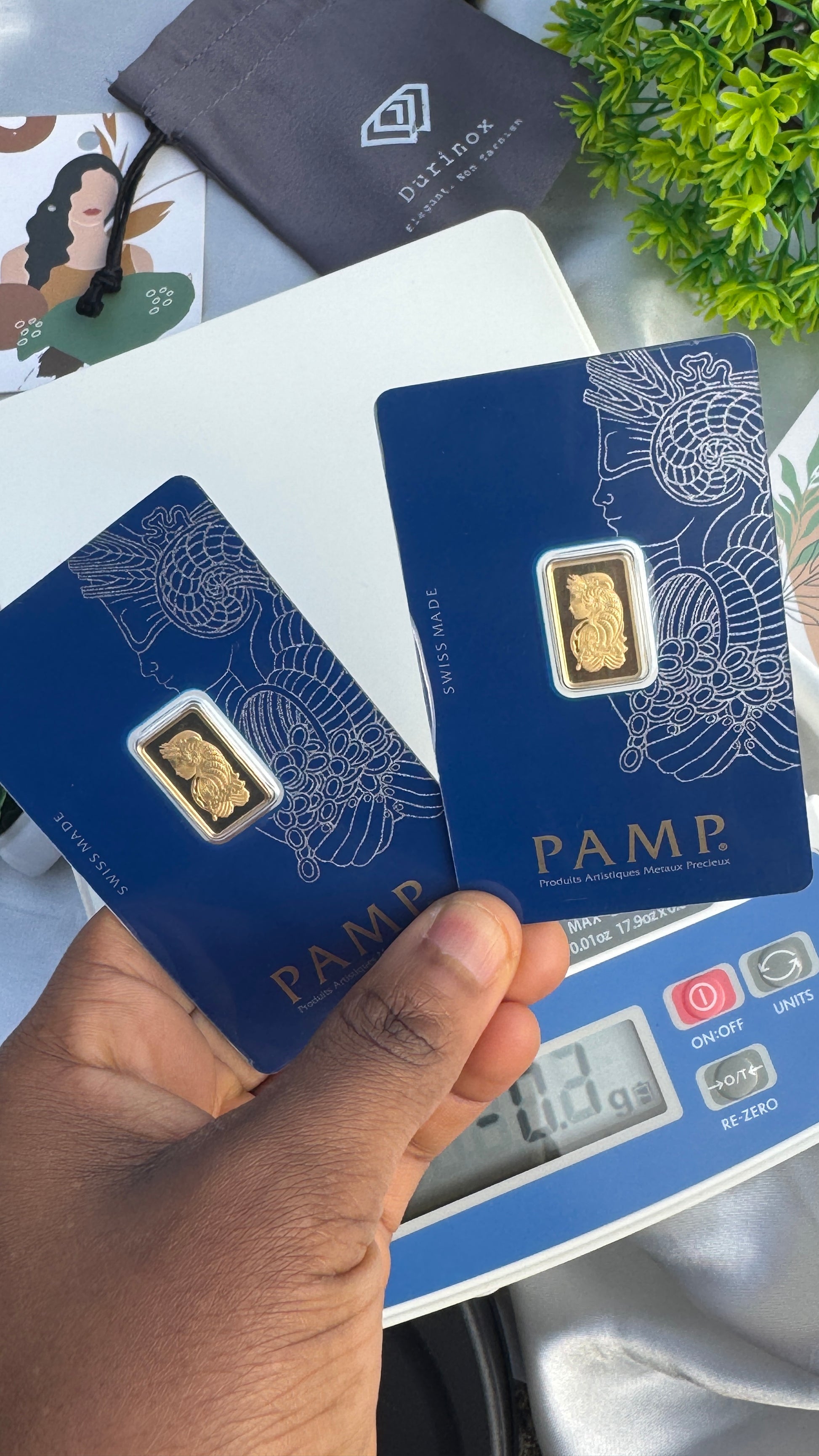 Gold bar on a PAMP card held by a hand with a digital scale in the background.