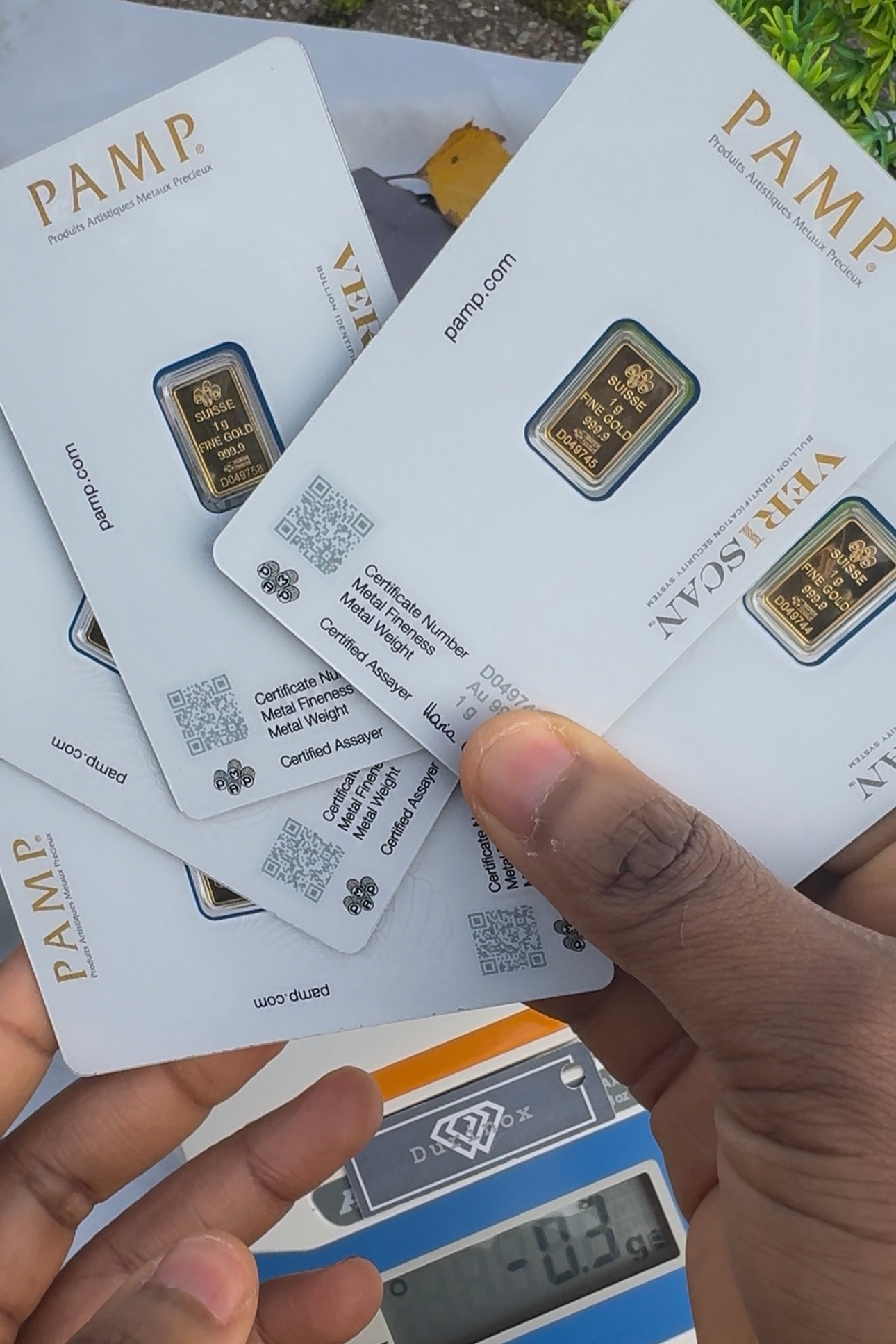 Gold bars in PAMP packaging held by a hand with a digital scale in the background.